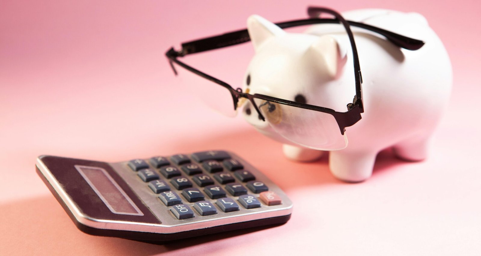Ein pinkes Sparschwein mit schwarzer Brille steht neben einem Taschenrechner auf einem weißen Hintergrund.