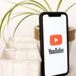 Ein Smartphone mit YouTube-Logo liegt auf einem Tisch neben einer kleinen Topfpflanze, Symbol für digitales Mediennutzung.
