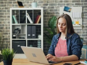 Eine Frau sitzt konzentriert an einem Schreibtisch und arbeitet an einem Laptop in einem modernen Büroraum.