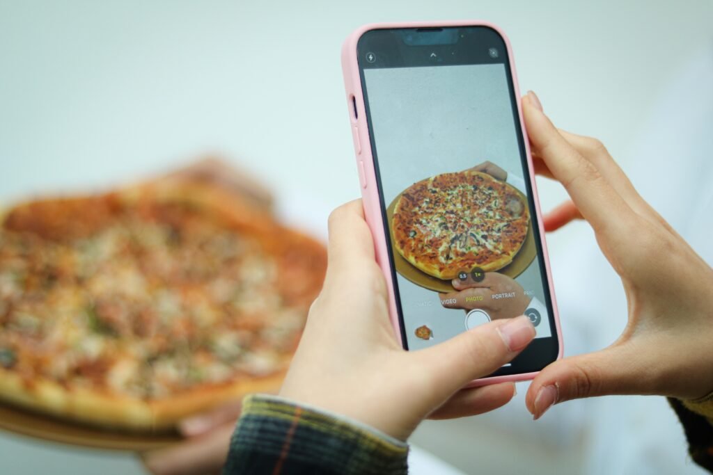 Eine Frau fotografiert eine Pizza mit ihrem Smartphone auf einem Tisch, Fokus auf das Essen und das Fotografieren.