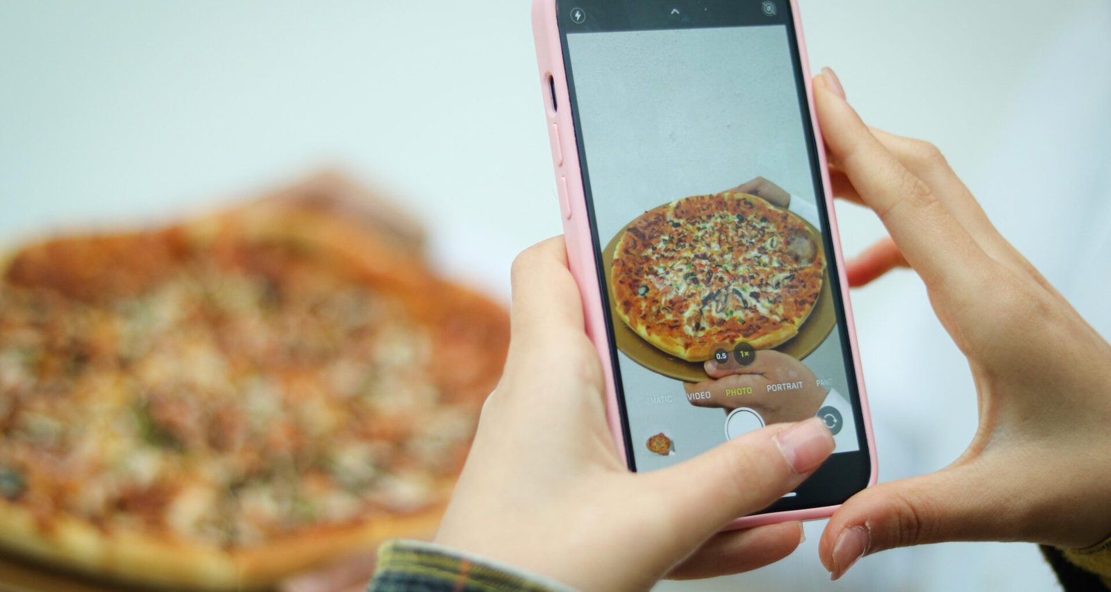 Eine Frau fotografiert eine Pizza mit ihrem Smartphone auf einem Tisch, Fokus auf das Essen und das Fotografieren.