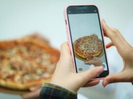 Eine Frau fotografiert eine Pizza mit ihrem Smartphone auf einem Tisch, Fokus auf das Essen und das Fotografieren.