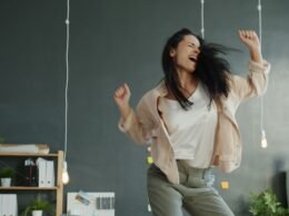 Eine Frau tanzt mit ausgestreckten Armen vor neutralem Hintergrund. Dynamische Pose, seitlicher Blick auf die Tänzerin.