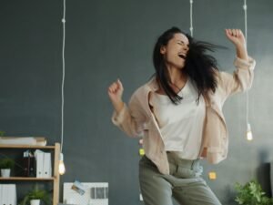 Frau tanzt fröhlich mit erhobenen Armen vor neutralem Hintergrund, ausdrucksstarke Körperbewegung eingefangen