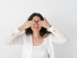 Frau sitzt vor hellem Hintergrund und bedeckt ihre Augen mit beiden Händen als Zeichen für Anonymität im Internet.