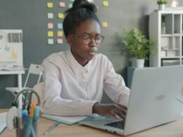 Eine Frau arbeitet an einem Laptop in einem modernen Büroraum mit Tageslicht und Schreibtisch.