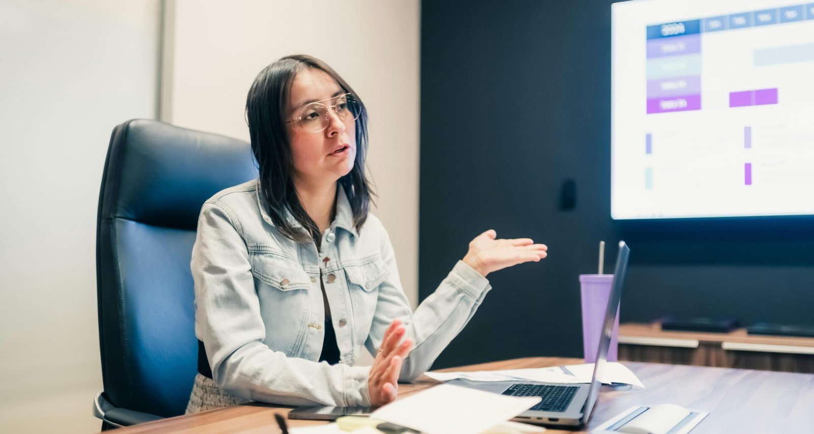 Frau steht vor einer Präsentationsleinwand und erklärt Inhalte gestikulierend während einer Besprechung.