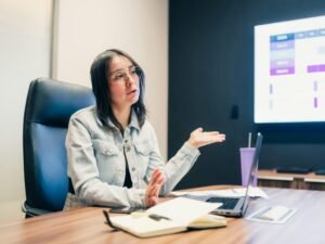 Frau steht vor einer Präsentationsleinwand und erklärt Inhalte gestikulierend während einer Besprechung.