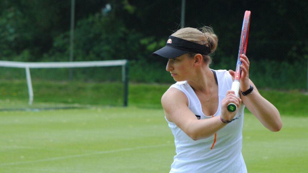 Eine Frau hält einen Tennisschläger und steht auf einem Tennisplatz, bereit für das Spiel. Sie trägt sportliche Kleidung.