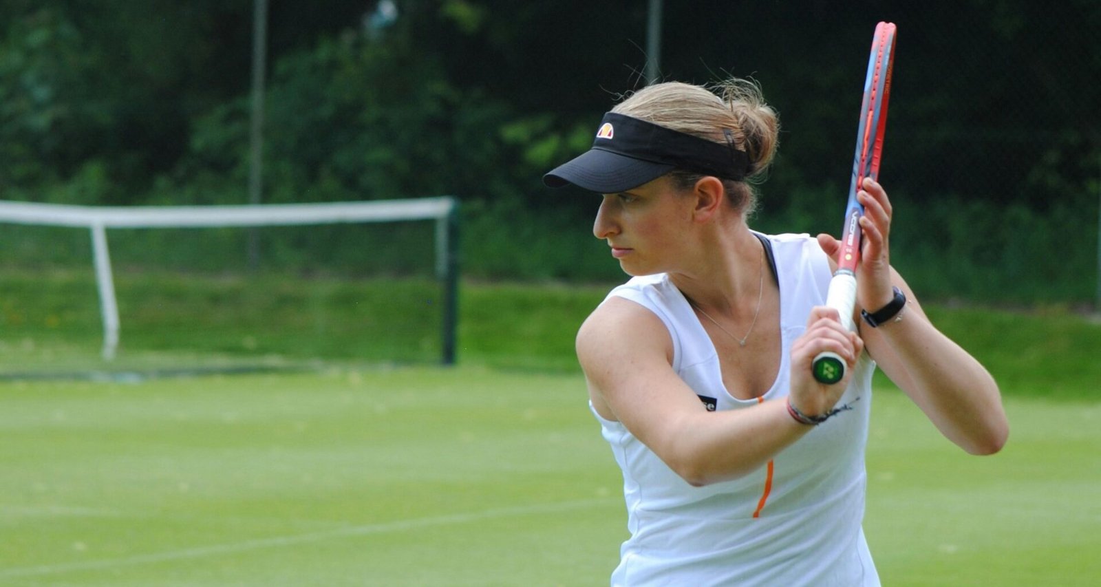Eine Frau hält einen Tennisschläger und steht auf einem Tennisplatz, bereit für das Spiel. Sie trägt sportliche Kleidung.