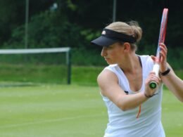 Eine Frau hält einen Tennisschläger und steht auf einem Tennisplatz, bereit für das Spiel. Sie trägt sportliche Kleidung.