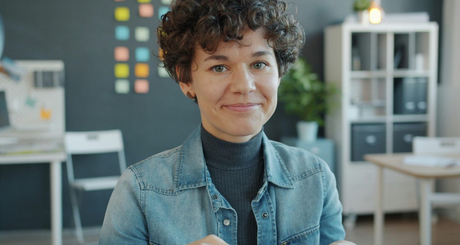 Frau mit lockigen Haaren sitzt lächelnd an einem Schreibtisch in einem hellen, modernen Büro