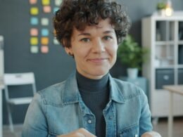 Frau mit lockigen Haaren sitzt lächelnd an einem Schreibtisch in einem hellen, modernen Büro