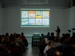 Viele Personen sitzen dicht beieinander in einem modernen, hellen Raum und nehmen an einer Veranstaltung teil.