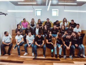 Mehrere Personen sitzen nebeneinander auf einer langen Holzbank im Freien und unterhalten sich miteinander.