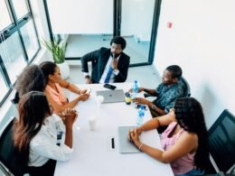 Eine Gruppe von Menschen sitzt um einen weißen Tisch und arbeitet gemeinsam an Laptops und Dokumenten.