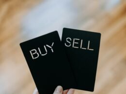 Eine Hand hält zwei schwarze Karten mit den Aufschriften 'buy' und 'sell', symbolisch für digitale Handelsprozesse