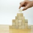 Eine menschliche Hand platziert ein Holzstück in eine Holzpyramide auf einer ebenen Fläche, Symbol für Organisation und Strategie.