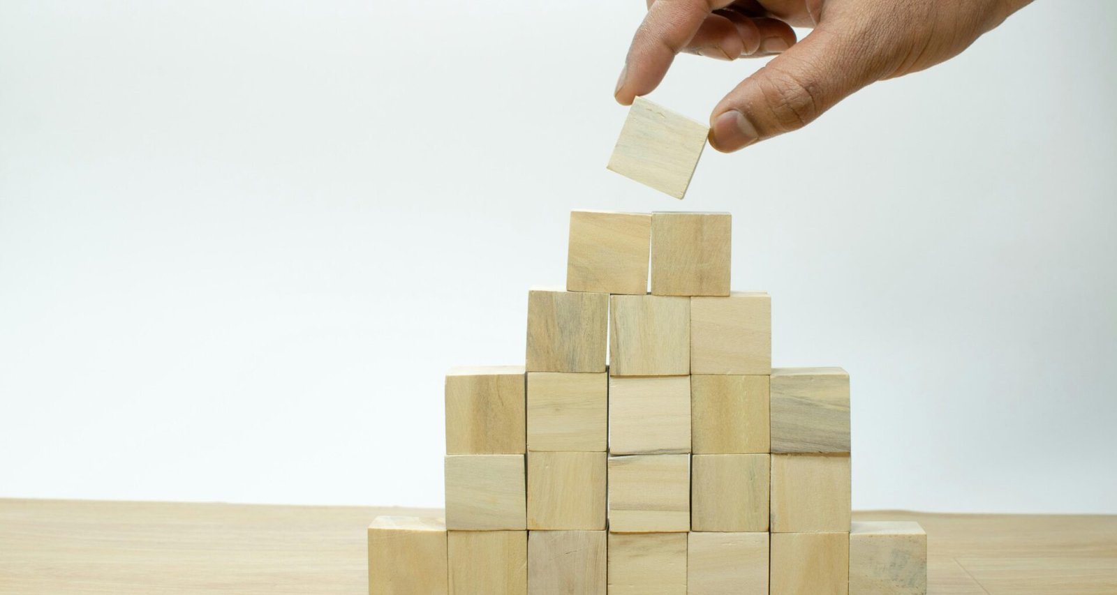Eine menschliche Hand platziert ein Holzstück in eine Holzpyramide auf einer ebenen Fläche, Symbol für Organisation und Strategie.