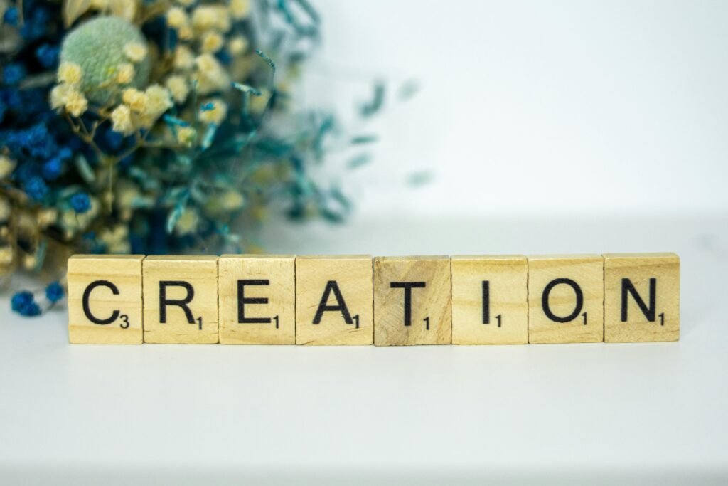 Ein Holzblock mit der Aufschrift Creation steht auf einer Oberfläche, im Hintergrund befinden sich Blumen.