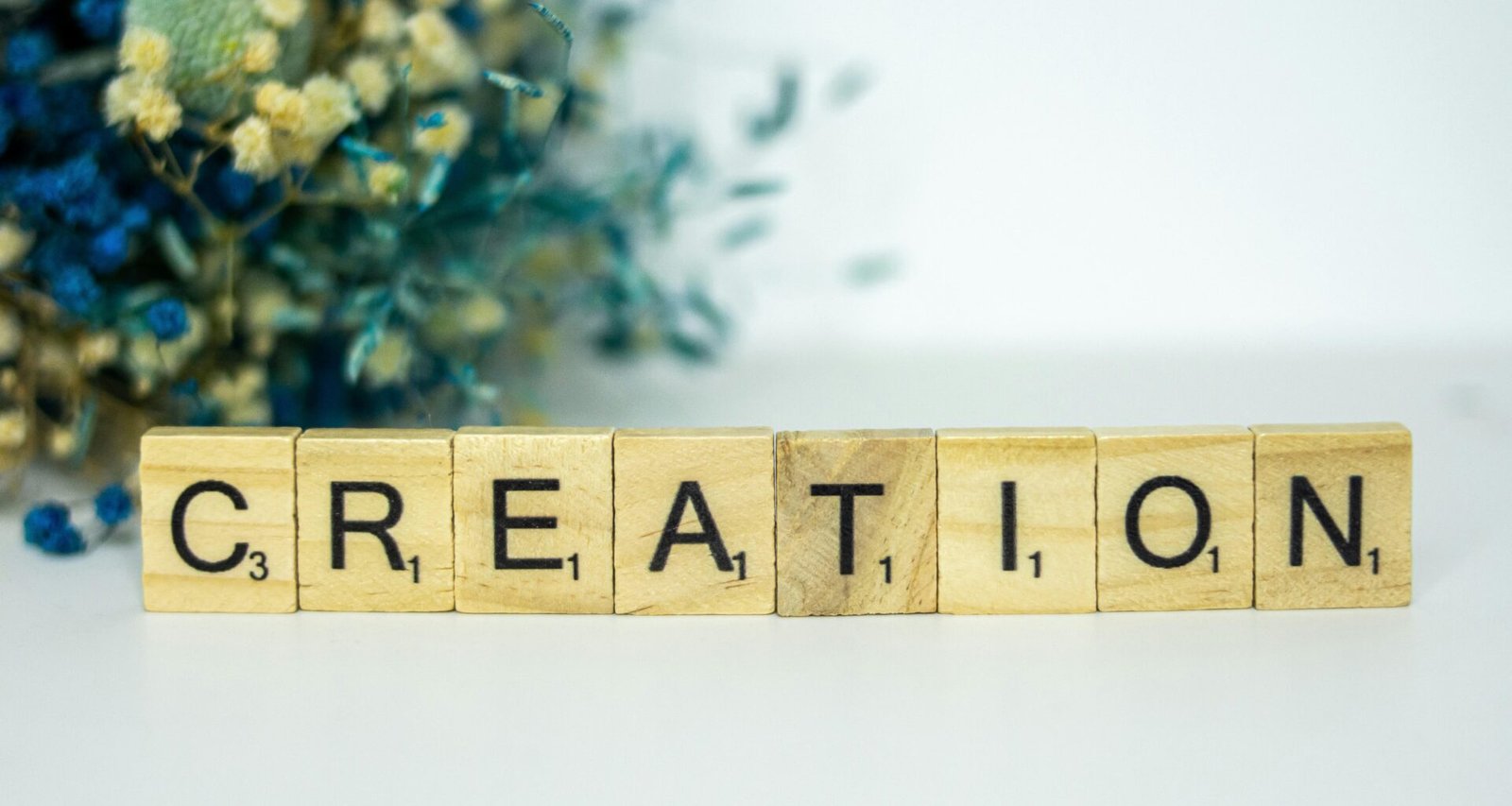 Ein Holzblock mit der Aufschrift Creation steht auf einer Oberfläche, im Hintergrund befinden sich Blumen.