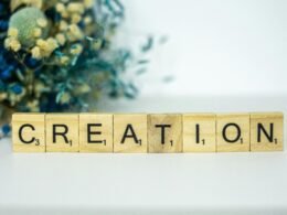 Ein Holzblock mit der Aufschrift Creation steht auf einer Oberfläche, im Hintergrund befinden sich Blumen.