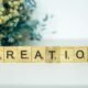 Ein Holzblock mit der Aufschrift Creation steht auf einer Oberfläche, im Hintergrund befinden sich Blumen.