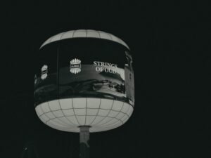 Ein beleuchtetes, kugelförmiges Werbeschild mit der Aufschrift 'strings of old' bei Nacht in urbaner Umgebung.