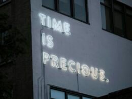 Leuchtendes Neon-Schild mit dem Text Time is precious auf dunklem Hintergrund, fotografiert von Jose Fabula