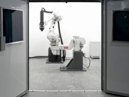 Eine Maschine steht zentral in einem technisch ausgestatteten Raum, verschiedene Leitungen und Geräte sind sichtbar.