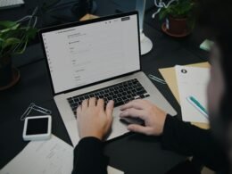 Eine Person sitzt an einem Schreibtisch und arbeitet an einem Laptop, umgeben von typischer Büroausstattung.