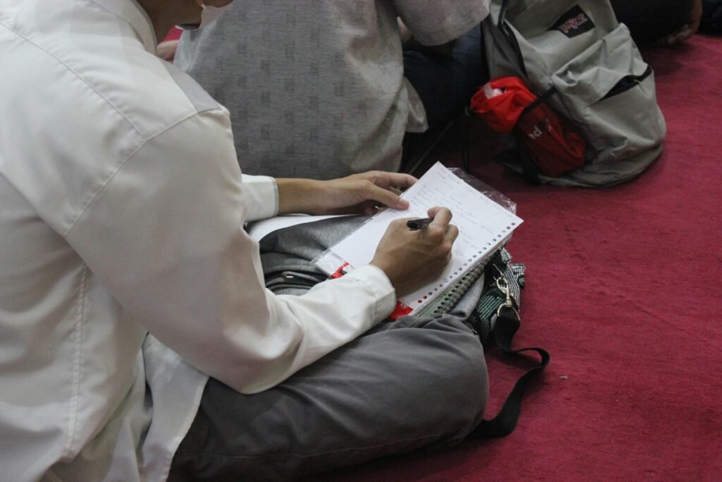 Eine Person sitzt an einem Tisch und schreibt mit einem Stift handschriftlich auf ein weißes Blatt Papier.