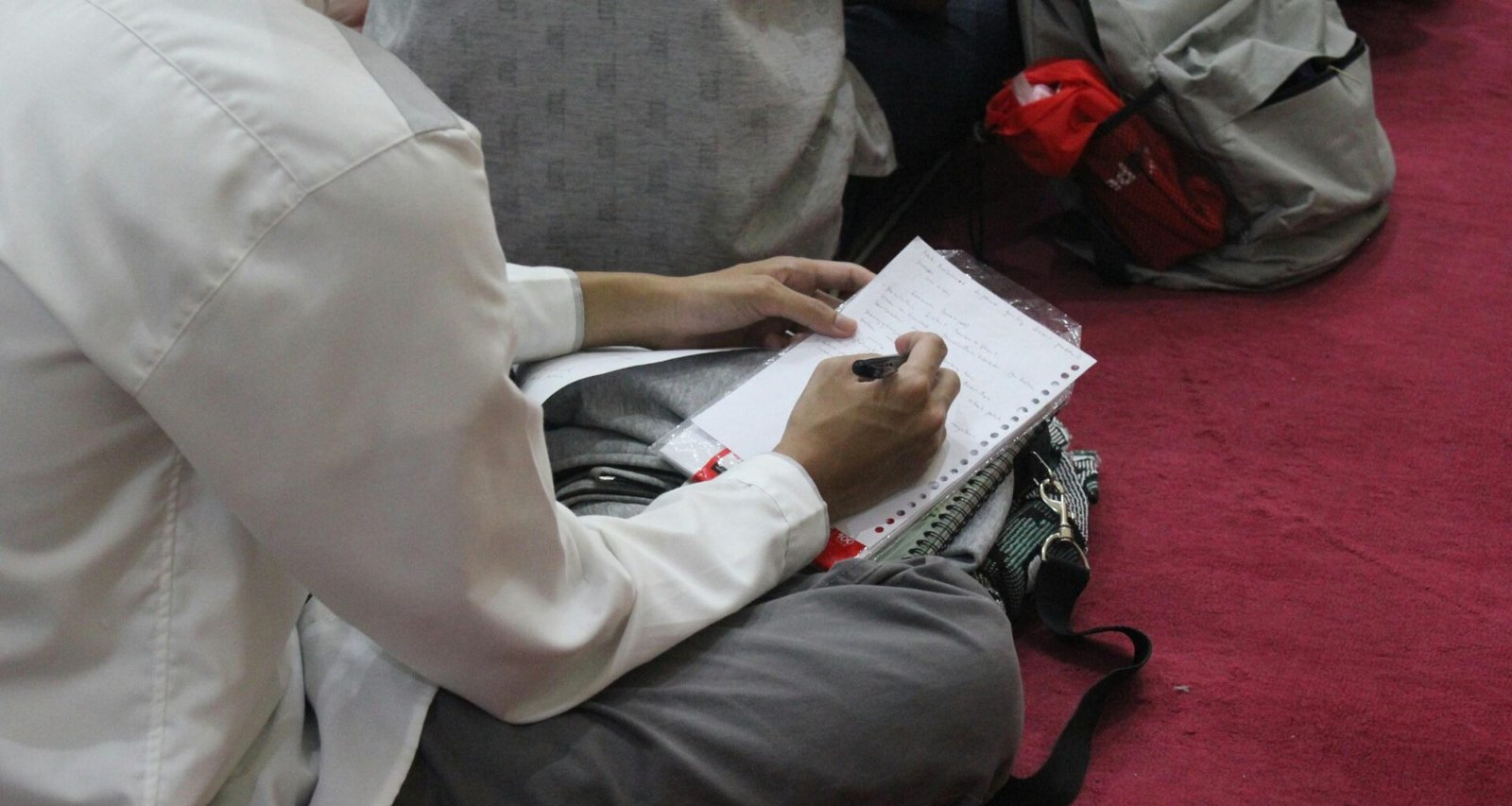 Eine Person sitzt an einem Tisch und schreibt mit einem Stift handschriftlich auf ein weißes Blatt Papier.