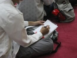 Eine Person sitzt an einem Tisch und schreibt mit einem Stift handschriftlich auf ein weißes Blatt Papier.