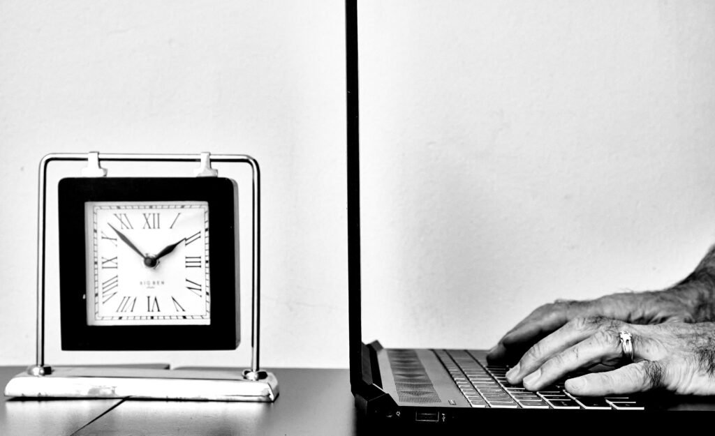 Person tippt auf einem Laptop neben einer analogen Uhr auf einem Tisch – Symbol für digitales Arbeiten und Zeitmanagement