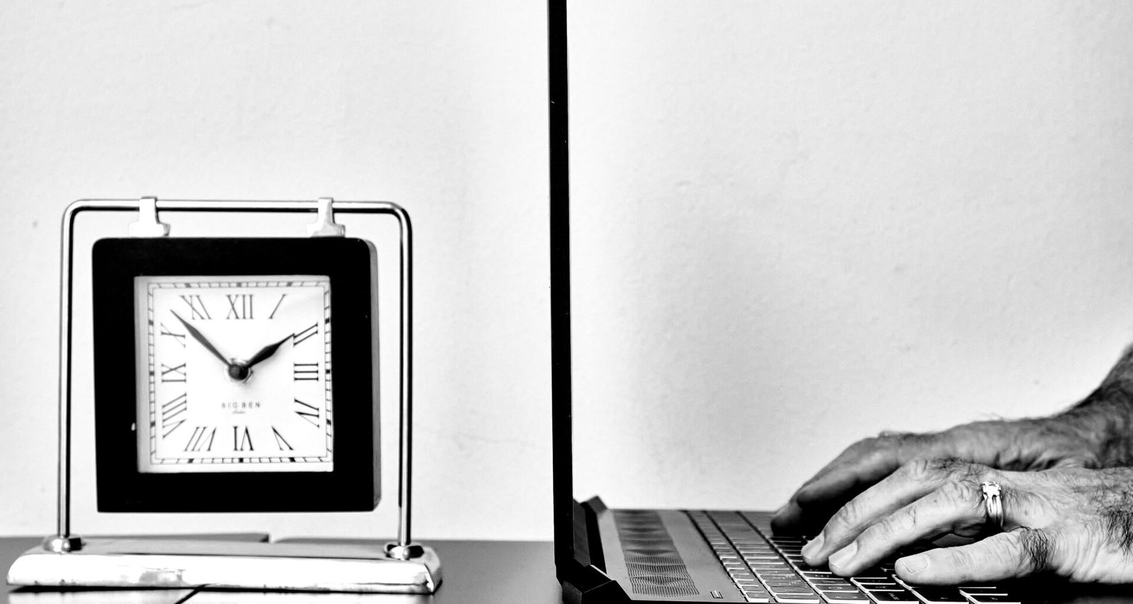 Eine Person tippt an einem Laptop auf einem Schreibtisch, neben ihr steht eine analoge Uhr und weiteres Arbeitszubehör.