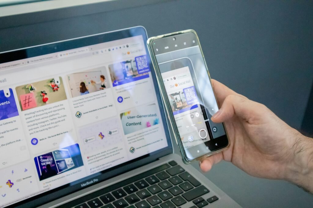 Eine Person hält ein Smartphone in der Hand und sitzt neben einem geöffneten Laptop an einem Tisch.