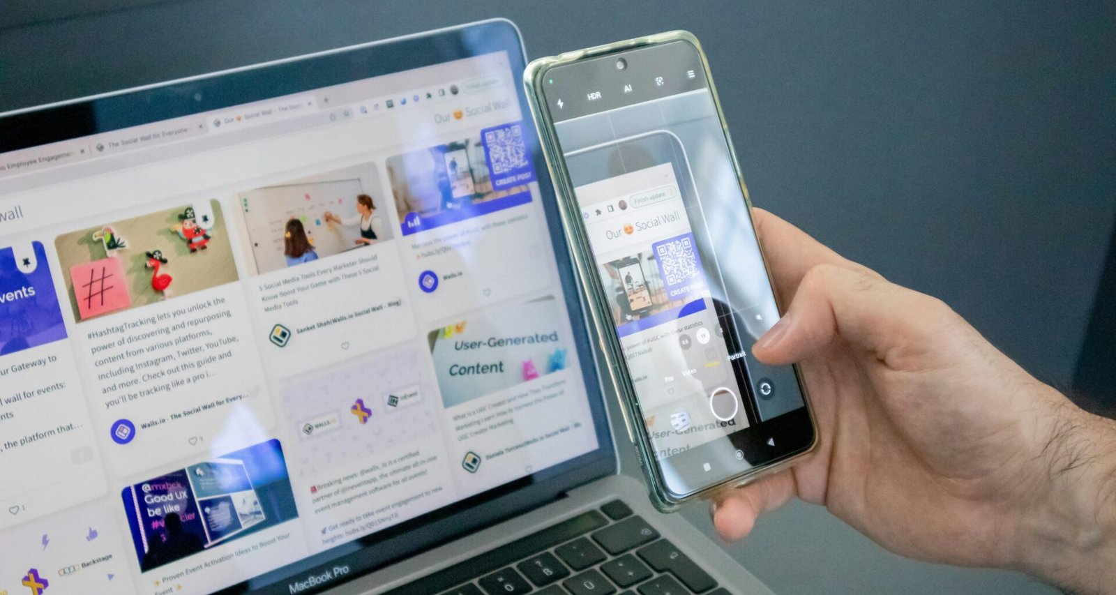 Eine Person hält ein Smartphone in der Hand und sitzt neben einem geöffneten Laptop an einem Tisch.