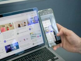 Eine Person hält ein Smartphone in der Hand und sitzt neben einem geöffneten Laptop an einem Tisch.