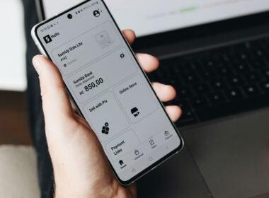 Eine Person hält ein Smartphone vor einem offenen Laptop, beide Geräte werden im Arbeitsalltag parallel genutzt.