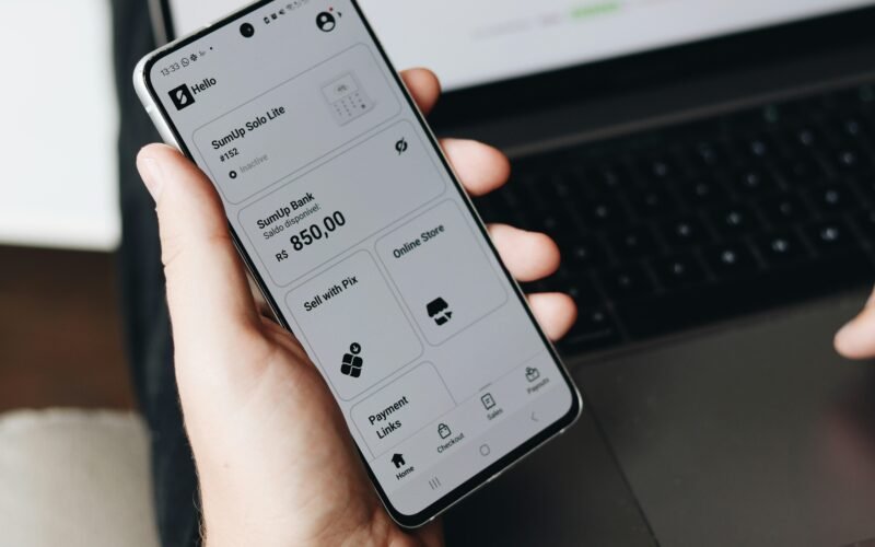 Eine Person hält ein Smartphone vor einem offenen Laptop, beide Geräte werden im Arbeitsalltag parallel genutzt.