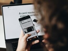 Person hält ein Smartphone vor einem aufgeklappten Laptop, beide Geräte sind im Arbeitsumfeld sichtbar.