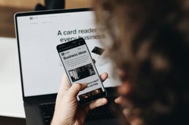 Person hält ein Smartphone vor einem aufgeklappten Laptop, beide Geräte sind im Arbeitsumfeld sichtbar.