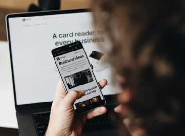 Person hält ein Smartphone vor einem aufgeklappten Laptop, beide Geräte sind im Arbeitsumfeld sichtbar.