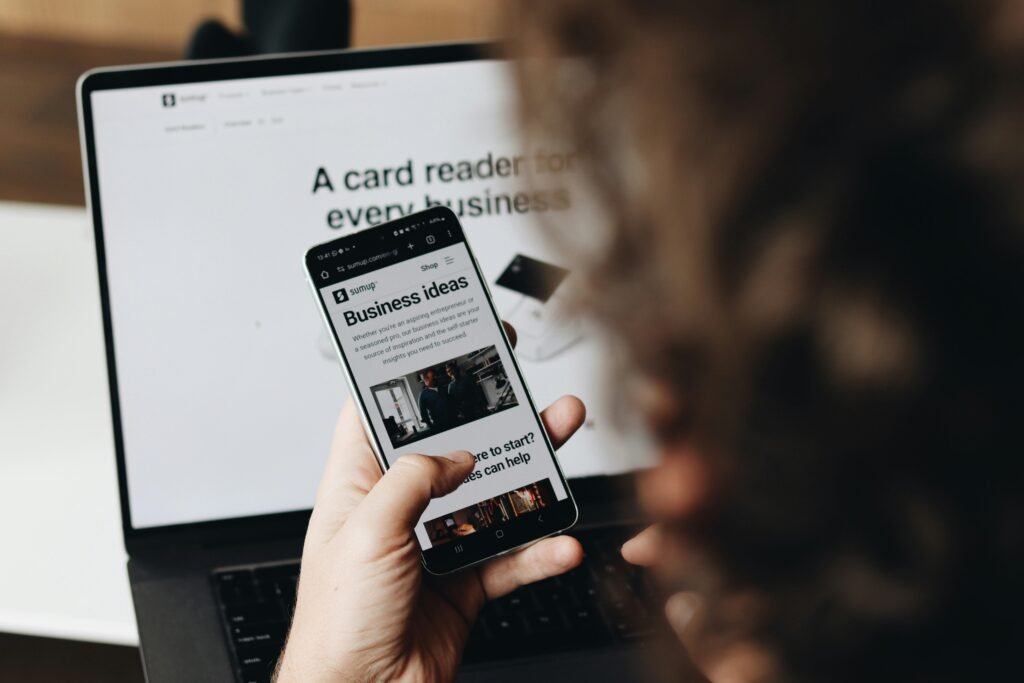 Eine Person hält ein Smartphone vor einem aufgeklappten Laptop an einem Arbeitsplatz.