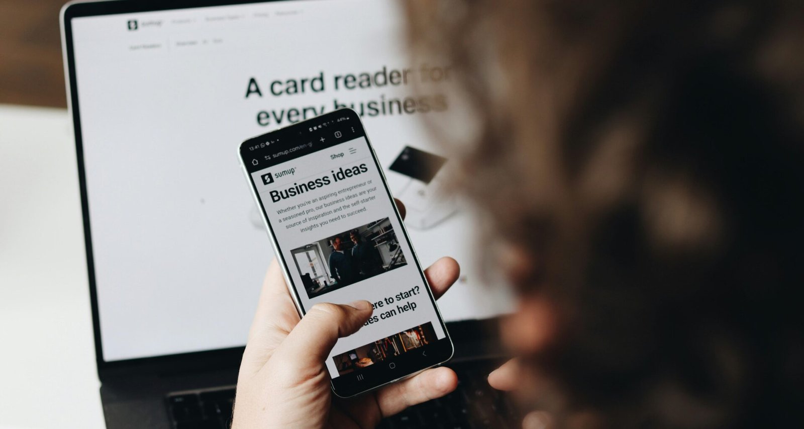 Eine Person hält ein Smartphone vor einem aufgeklappten Laptop an einem Arbeitsplatz.