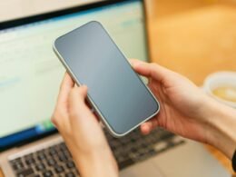 Person hält ein Smartphone in der Hand vor einem geöffneten Laptop auf einem Arbeitstisch, Fokus auf Mobile Nutzung.