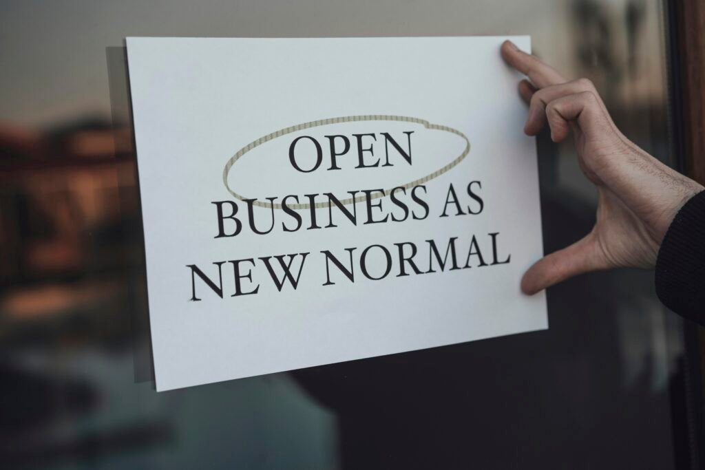 Eine Person hält ein Schild mit der Aufschrift ‚Open Business as New Normal‘ vor einem Geschäftseingang
