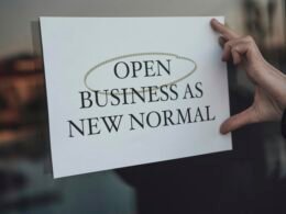 Eine Person hält ein Schild mit der Aufschrift ‚Open Business as New Normal‘ vor einem Geschäftseingang