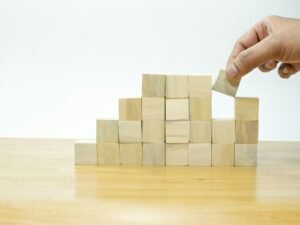 Eine Person legt einen Holzblock auf einen Stapel aus Holzklötzen, als Symbol für Strategie und Aufbau im digitalen Kontext.